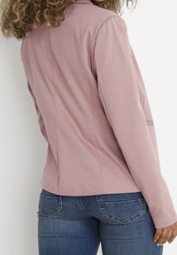 Giacca aderente di un rosa chiaro, con maniche lunghe, colletto e dettagli delle cuciture posteriori. Abbinata a jeans in denim scuro con tasche cucite.