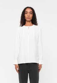 Witte blouse met lange mouwen, een ronde halslijn en een sleutelgatdetail aan de voorkant. Bevat een textuurstof en gathered schouders. Gepaard met donkere spijkerbroeken.