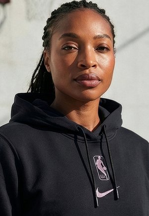Junge Frau mit geflochtenem Haar trägt einen schwarzen NBA Nike Hoodie und schaut selbstbewusst in die Kamera vor hellem Hintergrund.