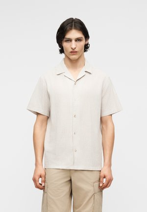 Mladi moški s srednje dolgimi temnimi lasmi, oblečen v svetlo bež srajco s kratkimi rokavi z gumbi in bež cargo kratke hlače, stoji pred enobarvnim ozadjem.