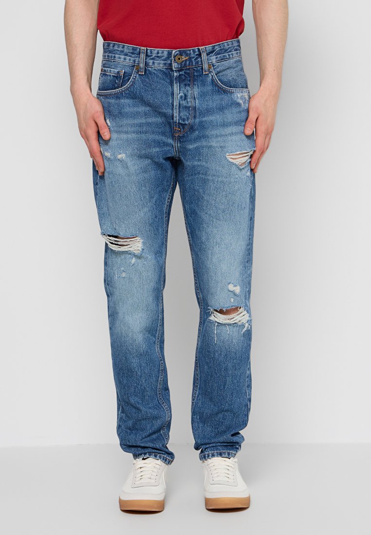 Ausgeblichene blaue Jeans aus Denim mit distanzierenden Rissen an den Knien, fünf Taschen und geradem Schnitt. Kombiniert mit weißen Sneakern.