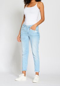 Světlé modré džíny s úzkým střih, se šesti kapsami, zapínáním na knoflík a mírně vybledlou texturou. Doplněno o bílé tílko a tenisky.