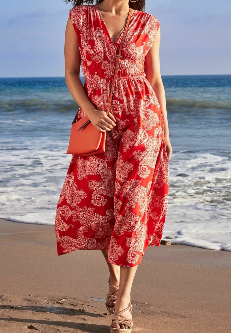 Femme portant une robe rouge à motif cachemire et des sandales beige, tenant un sac bandoulière orange, marchant sur une plage près de l'océan.