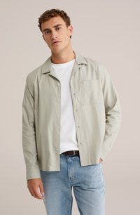 Svetlo zelena srajca s kratkimi rokavi in žepom na prsih, z dolgimi rokavi in klasičnim ovratnikom, oblečena čez belo majico in v kombinaciji z modrimi džinsami.