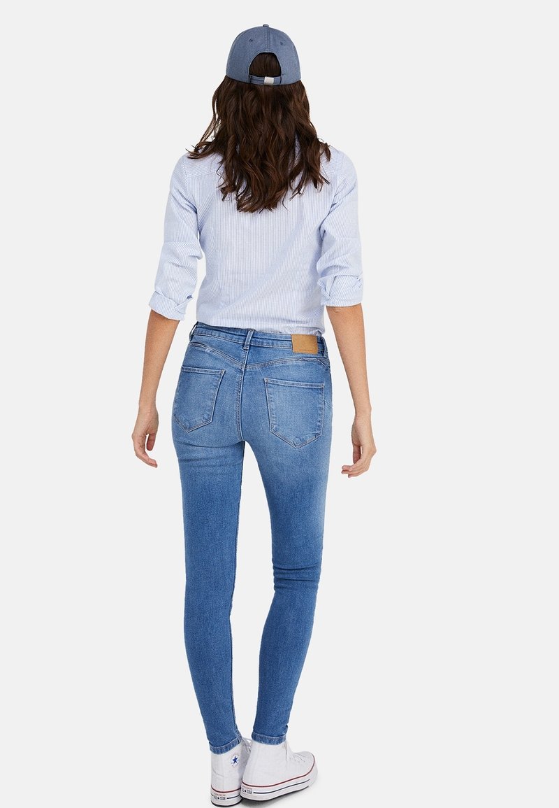 Jean skinny en denim bleu clair, avec des poches arrière et une étiquette en cuir marron. Associé à un haut à manches longues rayé et une casquette.
