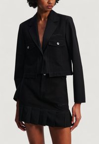 Giacca nera corta con due tasche frontali, hardware argentato e logo ricamato. Abbinata a una gonna coordinata con orlo plissettato.