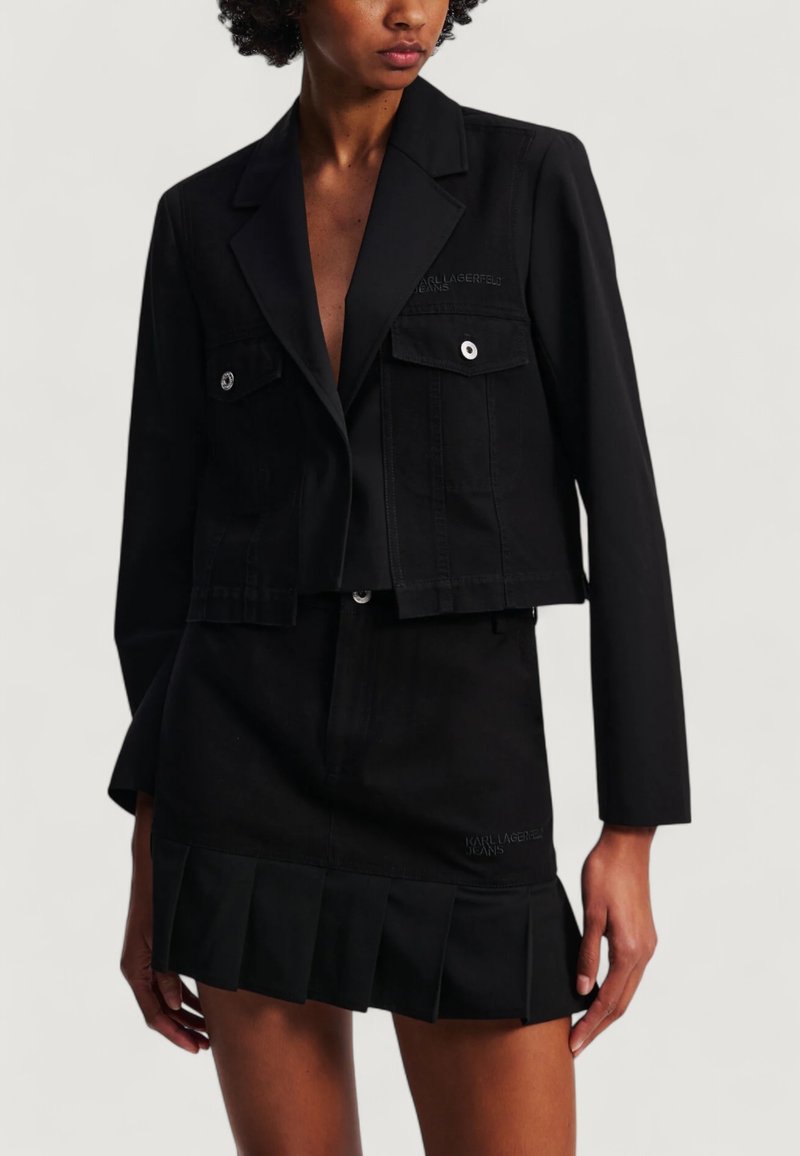 Giacca nera corta con due tasche frontali, hardware argentato e logo ricamato. Abbinata a una gonna coordinata con orlo plissettato.