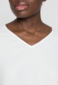 Hvid V-hals top med en glat tekstur, let stof og en subtil glans. Nærbillede der fremhæver halsudskæringen og hudtonen.