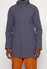 Homme portant un imperméable à capuche gris foncé longueur genou avec manches longues et pantalon orange, visible des épaules jusqu'à mi-cuisse.