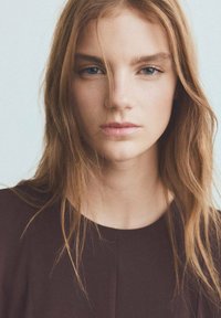 Portrait d'une personne aux longs cheveux châtains ondulés, aux yeux bleu clair, à la peau lisse, portant un haut foncé sur un fond clair.