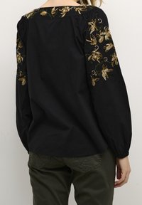 Blouse noire avec broderie florale en or et vert. Dotée de manches bouffantes et d'un col en U. Tissu doux avec une coupe décontractée.