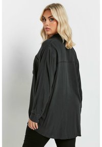 Camisa larga negra hecha de tejido suave, con un corte holgado, dobladillo ligeramente curvado y puños abotonados. Detalle de costura en la parte posterior.