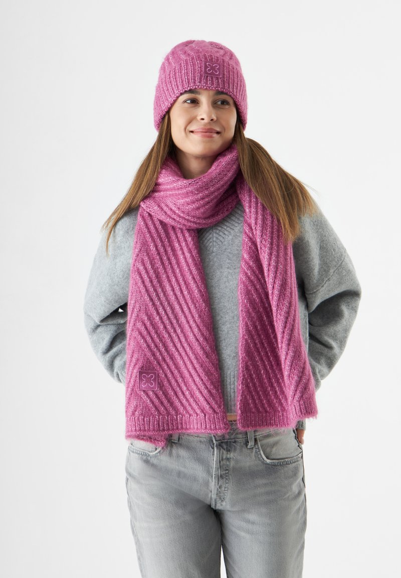 Roze gebreide muts en sjaal set met ribbeltextuur, voorzien van diagonale strepen. Gekozen boven een grijze trui; subtiele logo-afwerking op de accessoires.