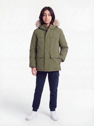 Teenager iført en olivengrøn parka med pelsforet hætte, marineblå cargobukser og hvide sneakers, stående mod en hvid baggrund.
