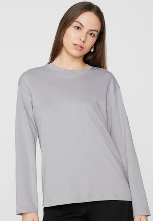 Młoda kobieta o długich brązowych włosach, ubrana w jasnoszarą bluzkę z długim rękawem i czarne spodnie, zwrócona przodem na tle jednolitego tła.