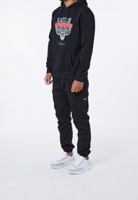 Sudadera negra con un gráfico de tigre y una franja roja, combinada con pantalones cargo negros y zapatillas blancas. Cuenta con un bolsillo frontal y puños acanalados.