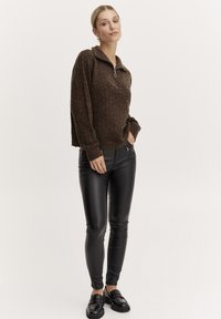 Brauner strukturierter Pullover mit hohem Kragen und halbem Reißverschluss, kombiniert mit schmal geschnittenen schwarzen Kunstlederhosen und schwarzen Slippern.