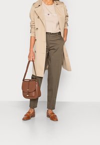 Beige trenchcoat, ljusbrun t-shirt med logotyp, olivgröna byxor, brun läderväska och matchande loafers. Släta texturer tydliga.