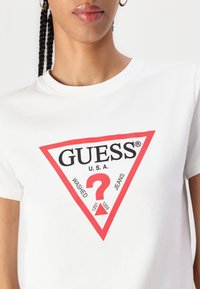T-shirt blanc à manches courtes présentant un design de triangle rouge avec le texte noir "GUESS", "U.S.A." et "WASHED JEANS", accompagné d'un graphique de point d'interrogation central.
