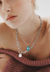 Collares en capas de plata con una cadena gruesa y un colgante ovalado de turquesa con un diseño, llevados con una prenda de punto burdeos.
