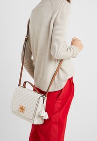Donna che indossa un maglione a coste beige e pantaloni rossi, mentre porta una borsa a tracolla bianca con motivo e una tracolla marrone con dettaglio a frange.