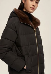 Giacca puffa nera con colletto in pelliccia sintetica marrone, zip dorata e tasche con bottoni a scatto. Texture morbida e design trapuntato.