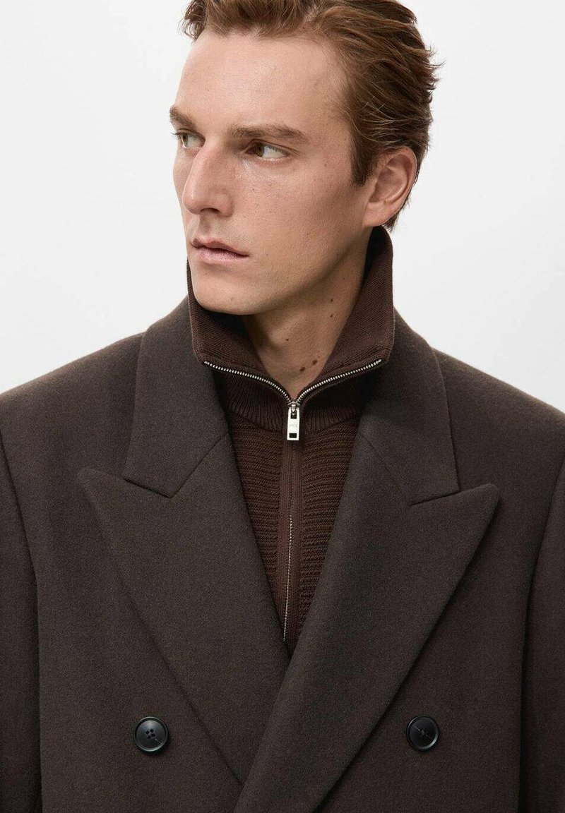 Cappotto doppio petto in lana marrone con collo alto, caratterizzato da ampi revers e uno strato interno con cerniera di un marrone più scuro con una texture a coste.