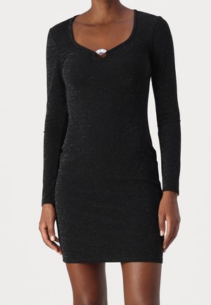 Mujer que lleva un vestido mini negro ajustado de manga larga con un sutil brillo y un pequeño detalle de ojo de cerradura cerca del escote.