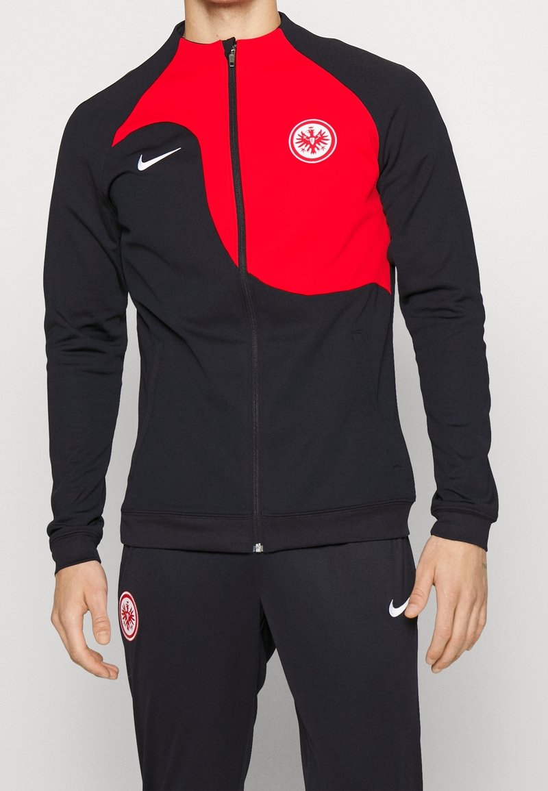 Chaqueta de chándal Nike en negro y rojo, con cierre de cremallera, mangas largas y acentos de logo. Tejido suave con un diseño en bloques de color.