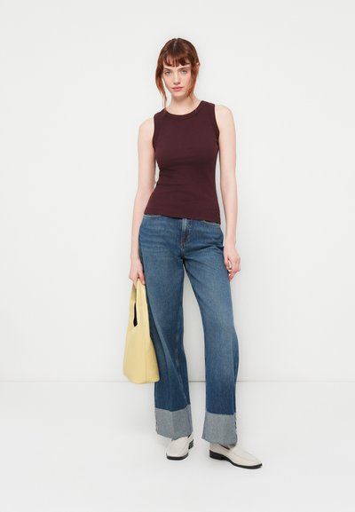 Top sans manches bordeaux foncé, jeans bleu à larges jambes avec des poignets en denim clair, sac épaule beige et bottines crème sur un fond uni.