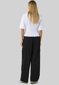 Bijela skraćena majica s teksturiranim leđima, u kombinaciji s labavim crnim cargo hlačama. Naglašena zelenim cipelama, prikazana s leđa na sivoj pozadini.