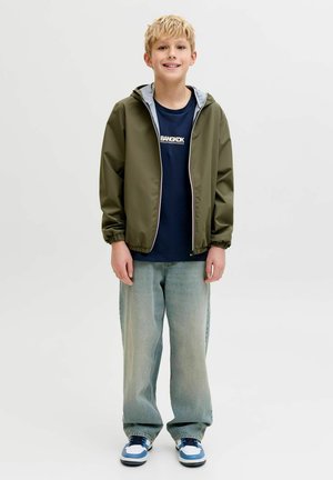 Šviesiaplaukis berniukas stovintis, dėvintis alyvuogių spalvos gobtuvu megztą striukę, tamsiai mėlyną "Bangkok" marškinėlį, laisvus šviesiai mėlynus džinsus ir baltus bei mėlynus sportinius batelius.