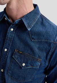 Artimo vaizdas asmens, dėvinčio mėlyną džinsinę „Lee“ marškinių su baltomis snap sagomis ir krūtinės kišenėle ant šviesiai pilkos fono.