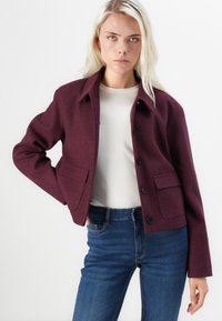 Burgundy wool jacket with a collared neckline, button front, and two front patch pockets, paired with a cream top and blue jeans.