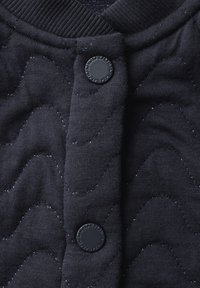Haut en tissu matelassé bleu marine avec un motif ondulé texturé, des boutons en plastique noirs et un col côtelé. Texture douce avec un léger éclat.