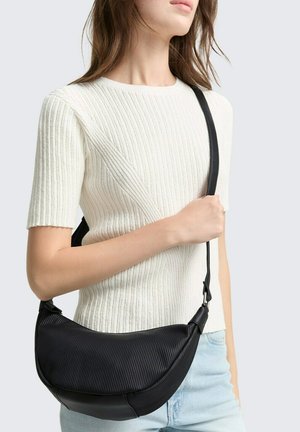 Schwarze gerippte Crossbody-Tasche mit geschwungener Form und verstellbarem Tragegurt, getragen über einem cremefarbenen gerippten Pullover, der Textur und Design hervorhebt.