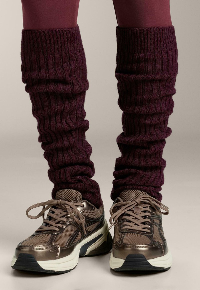 Ciorapi de gamă violetă cu textura ribbed se află deasupra unei perechi de adidași metalici de bronz cu detalii din plasă și tălpi albe, având șireturi și materiale texturate.