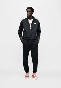 Μαύρ η αθλητική φόρμα με ζακέτα με φερμουάρ και παντελόνια με ελαστική μέση, σετ με κόκκινα πάνινα παπούτσια με λευκές σόλες και κορδόνια.