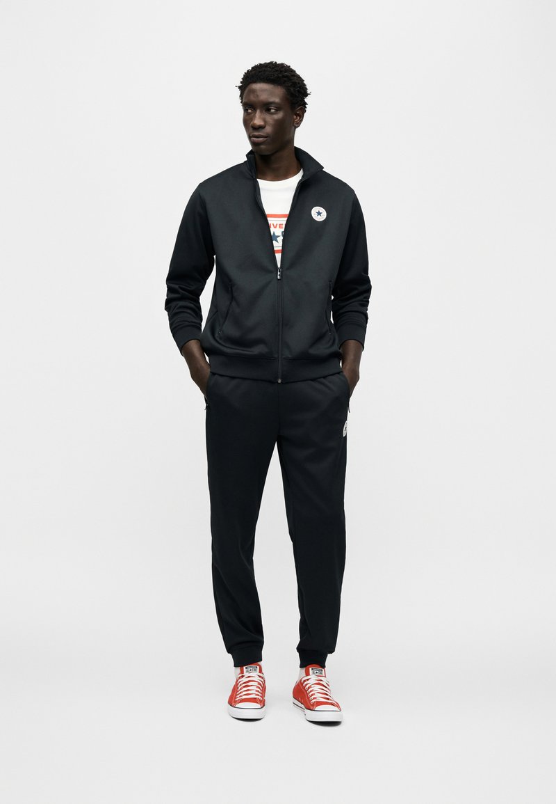 Schwarzer Sportanzug mit Reißverschlussjacke und Hosen mit elastischem Bund, kombiniert mit roten Leinenschuhen mit weißen Sohlen und Schnürsenkeln.