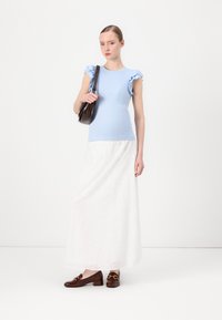 Haut côtelé bleu clair avec des manches volantes, jupe maxi blanche brodée, et mocassins marron avec des détails dorés, tenant un sac à main marron.