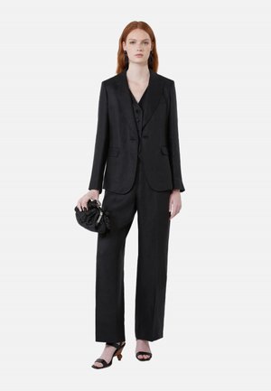 Donna rossa con un completo nero su misura, che tiene una pochette nera testurizzata e indossa sandali neri con tacco, in piedi davanti a uno sfondo bianco.