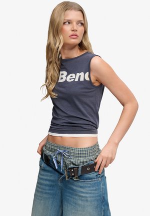 Ung kvinde med langt blondt hår iført en ærmeløs navy top med teksten "Bench", ternede shorts og løse blå jeans med sort bælte.