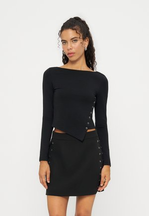 Woman wearing a black long-sleeve top with asymmetrical hem and matching black mini skirt, both with silver eyelet details on sides.