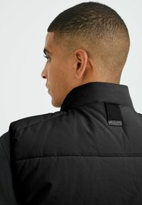 Crni puffer prsluk s visokim ovratnikom, teksturiranim materijalom i etiketom na leđima. Dizajn je elegantan s minimalnim hardverom.