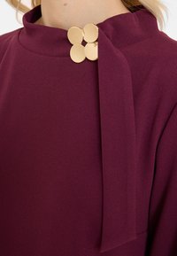 Close-up of a burgundy garment with a high collar fastened by four gold circular buttons on the shoulder.