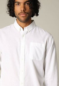 Chemise blanche à boutons faite d'un tissu léger, dotée d'une poche sur la poitrine et d'un col classique. La texture semble lisse et nette.