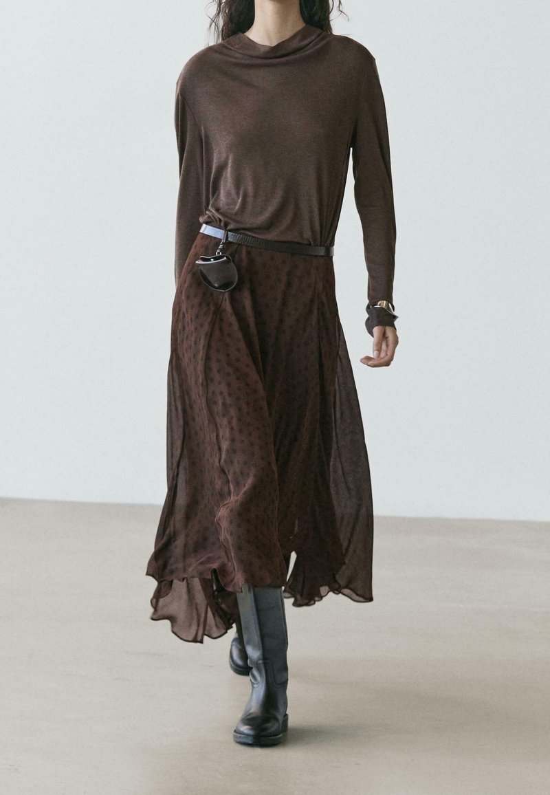 Femme marchant avec un haut marron à manches longues et une jupe fluide marron foncé à pois, des bottes noires montantes et un sac ceinture sur un sol clair.