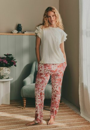 Frau mit lockigem blondem Haar, die ein cremefarbenes Oberteil mit Rüschenärmeln und rote Blumenhose trägt, steht barfuß in einem gemütlichen Wohnzimmer mit natürlichem Licht.