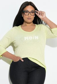 Jasnozielona, lekka koszula z długim rękawem, dekoltem w serek i napisem "MOIN". Połączona z ciemnymi dżinsami, które mają kieszeń po lewej stronie.