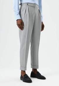 Pantalon gris à plis avec deux fermetures à boutons, ourlets retroussés et tissu texturé, associé à des mocassins en daim foncé.